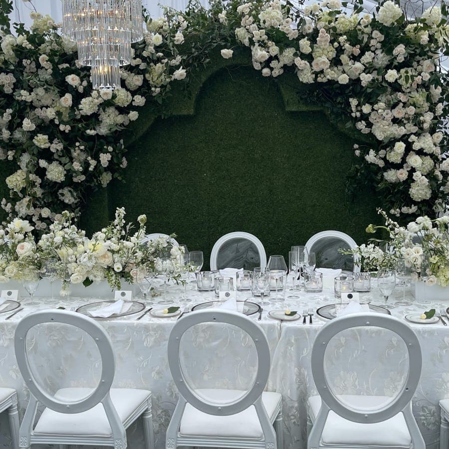 Reception table set with Louis-style chairs, glassware and low florals in front of a floral wall